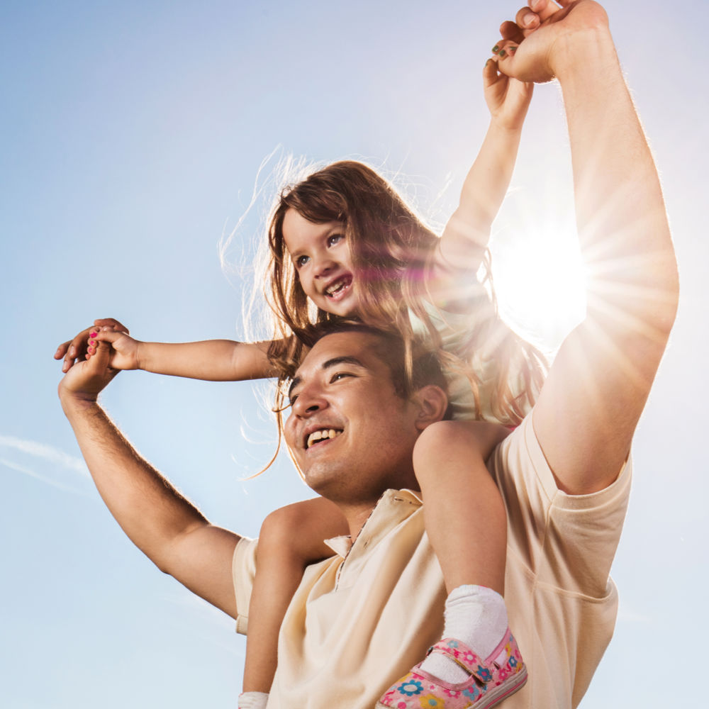 A smiling man with a young girl on his shoulders, holding hands, with a brilliant sun and a bright blue sky behind them